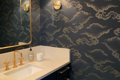 Small trendy dark wood floor and wallpaper powder room photo with shaker cabinets, blue cabinets, a one-piece toilet, blue walls, an undermount sink, quartz countertops, white countertops and a built-in vanity