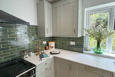 Photo of a medium sized classic l-shaped kitchen/diner in Hampshire with a belfast sink, shaker cabinets, green cabinets, quartz worktops, green splashback, ceramic splashback, black appliances, laminate floors, no island, brown floors and white worktops.
