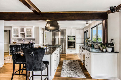 Elegant l-shaped medium tone wood floor and multicolored floor eat-in kitchen photo in Seattle with a farmhouse sink, recessed-panel cabinets, white cabinets, granite countertops, gray backsplash, porcelain backsplash, stainless steel appliances, an island and black countertops