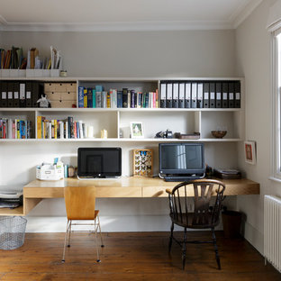 Foto på ett mellanstort funkis hemmabibliotek, med vita väggar, mellanmörkt trägolv, ett inbyggt skrivbord och brunt golv