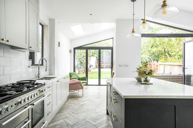 Photo of a transitional l-shaped kitchen in London with a double-bowl sink, shaker cabinets, white cabinets, white splashback, stainless steel appliances, medium hardwood floors, with island, brown floor and white benchtop.