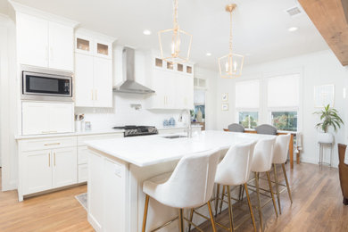 This is an example of a large traditional l-shaped open plan kitchen in New Orleans with a submerged sink, shaker cabinets, white cabinets, quartz worktops, white splashback, porcelain splashback, stainless steel appliances, light hardwood flooring, an island, brown floors and white worktops.