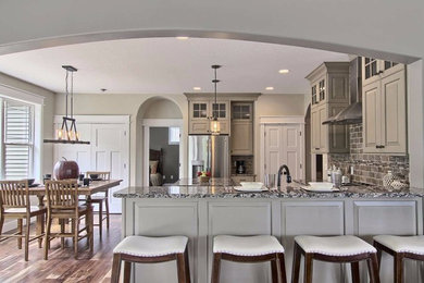 Example of a mid-sized mountain style u-shaped medium tone wood floor open concept kitchen design in Other with an undermount sink, raised-panel cabinets, beige cabinets, granite countertops, brown backsplash, stone tile backsplash, stainless steel appliances and an island