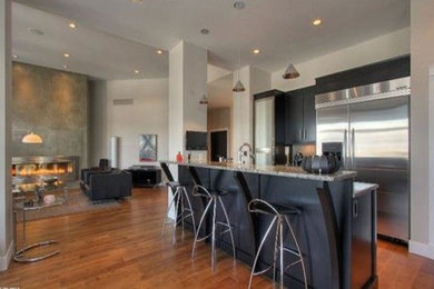 Mid-sized minimalist l-shaped light wood floor eat-in kitchen photo in Other with a farmhouse sink, flat-panel cabinets, black cabinets, granite countertops, gray backsplash, ceramic backsplash, stainless steel appliances and an island
