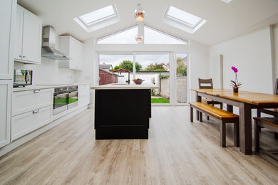 Photo of a mid-sized contemporary galley eat-in kitchen in Dublin with a drop-in sink, louvered cabinets, white cabinets, laminate benchtops, white splashback, black appliances, laminate floors, a peninsula, brown floor and white benchtop.