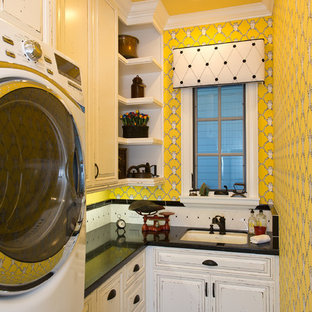 オレンジカウンティの中サイズのトラディショナルスタイルのおしゃれな洗濯室 (アンダーカウンターシンク、レイズドパネル扉のキャビネット、ヴィンテージ仕上げキャビネット、黄色い壁、上下配置の洗濯機・乾燥機、クオーツストーンカウンター、ライムストーンの床、黒い床、黒いキッチンカウンター) の写真
