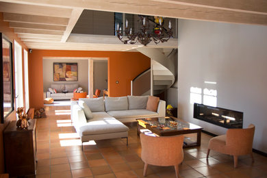 Réalisation d'un grand salon mansardé ou avec mezzanine design avec un mur blanc, tomettes au sol, une cheminée ribbon, un manteau de cheminée en plâtre et un sol orange.