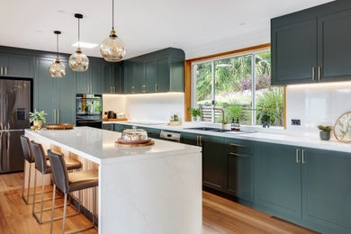 Large transitional l-shaped eat-in kitchen in Sydney with a drop-in sink, shaker cabinets, green cabinets, quartz benchtops, white splashback, engineered quartz splashback, black appliances, medium hardwood floors, with island, multi-coloured floor and white benchtop.