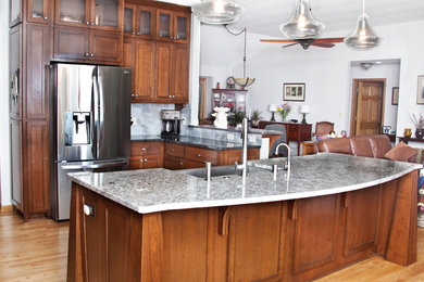 Open concept kitchen - transitional l-shaped light wood floor open concept kitchen idea in Milwaukee with a farmhouse sink, shaker cabinets, medium tone wood cabinets, granite countertops, gray backsplash, glass tile backsplash, stainless steel appliances and an island