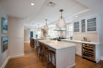 Example of a large transitional galley medium tone wood floor and brown floor open concept kitchen design in Boston with a farmhouse sink, shaker cabinets, white cabinets, marble countertops, gray backsplash, subway tile backsplash, stainless steel appliances, two islands and multicolored countertops