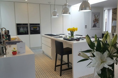This is an example of a mid-sized modern eat-in kitchen in London with a drop-in sink, flat-panel cabinets, white cabinets, quartzite benchtops, white splashback, mosaic tile splashback, stainless steel appliances, porcelain floors, with island, grey floor and white benchtop.