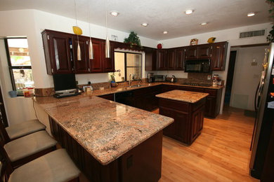 Elegant u-shaped light wood floor and brown floor enclosed kitchen photo in Phoenix with an undermount sink, raised-panel cabinets, dark wood cabinets, granite countertops, beige backsplash, stone slab backsplash, black appliances and two islands