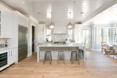 Large beach style u-shaped open plan kitchen in Charleston with light hardwood floors, with island, a farmhouse sink, shaker cabinets, white cabinets, marble benchtops, multi-coloured splashback, marble splashback, stainless steel appliances and beige floor.