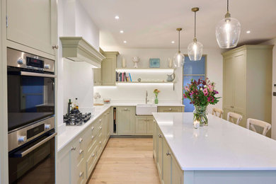 Medium sized classic l-shaped kitchen/diner in London with a belfast sink, shaker cabinets, green cabinets, quartz worktops, white splashback, black appliances, light hardwood flooring, an island, brown floors and white worktops.