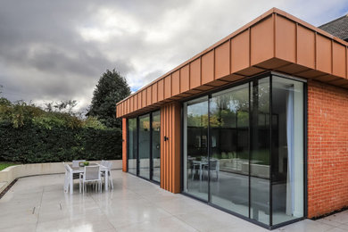 This is an example of a medium sized contemporary bungalow rear house exterior in Hampshire with metal cladding, an orange house, a flat roof, a mixed material roof and a grey roof.
