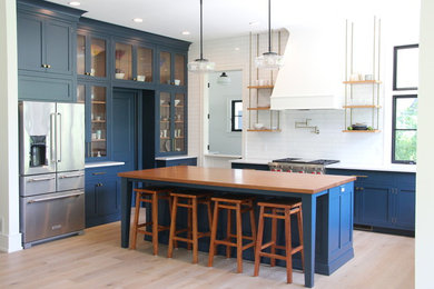 Mid-sized cottage l-shaped light wood floor and brown floor kitchen photo in Other with a farmhouse sink, shaker cabinets, blue cabinets, quartz countertops, white backsplash, subway tile backsplash, stainless steel appliances and an island