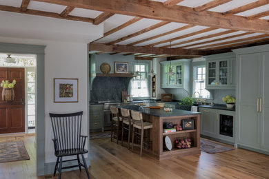 Example of a mid-sized country medium tone wood floor, brown floor and exposed beam eat-in kitchen design in Boston with a farmhouse sink, recessed-panel cabinets, green cabinets, soapstone countertops, black backsplash, stone slab backsplash, stainless steel appliances, an island and black countertops
