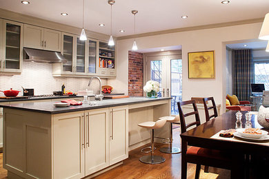 Mid-sized elegant l-shaped medium tone wood floor eat-in kitchen photo in New York with a farmhouse sink, shaker cabinets, beige cabinets, white backsplash, glass tile backsplash, stainless steel appliances and an island