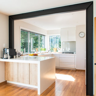 Mittelgroße Nordische Küche in U-Form mit Vorratsschrank, Unterbauwaschbecken, flächenbündigen Schrankfronten, weißen Schränken, Mineralwerkstoff-Arbeitsplatte, Glasrückwand, weißen Elektrogeräten, braunem Holzboden, Halbinsel und braunem Boden in Christchurch