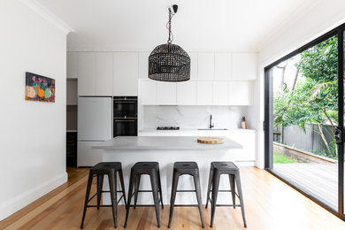This is an example of a contemporary kitchen in Sydney with an undermount sink, flat-panel cabinets, white splashback, stone slab splashback, white appliances, light hardwood floors, with island, beige floor and white benchtop.