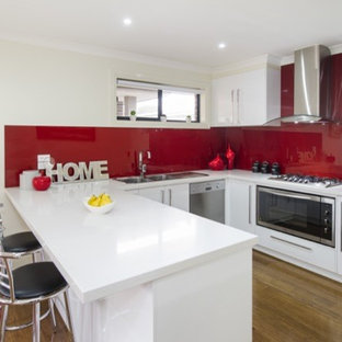 Cette image montre une arrière-cuisine design en U de taille moyenne avec un évier 2 bacs, un placard avec porte à panneau encastré, des portes de placard blanches, un plan de travail en stratifié, une crédence rouge, une crédence en feuille de verre, un électroménager en acier inoxydable, un sol en bambou et un îlot central.