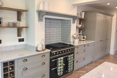 Inspiration for a mid-sized country galley eat-in kitchen in Sussex with a farmhouse sink, shaker cabinets, grey cabinets, quartzite benchtops, white splashback, ceramic splashback, black appliances, light hardwood floors, a peninsula, brown floor and grey benchtop.