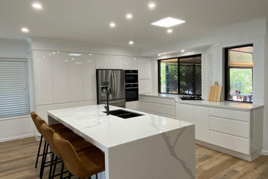 This is an example of a large modern l-shaped kitchen in Brisbane with an undermount sink, white cabinets, quartz benchtops, white splashback, subway tile splashback, black appliances, vinyl floors, with island, brown floor and white benchtop.