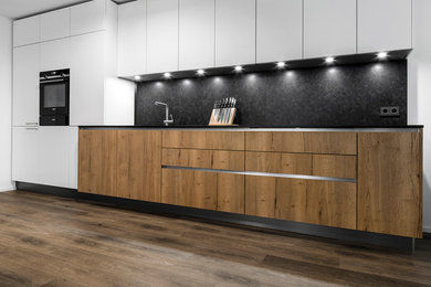 Design ideas for a mid-sized contemporary single-wall eat-in kitchen in Munich with a single-bowl sink, flat-panel cabinets, white cabinets, granite benchtops, grey splashback, stone slab splashback, black appliances, medium hardwood floors, no island, brown floor and grey benchtop.