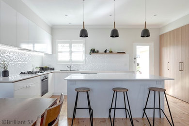 Inspiration for a mid-sized contemporary l-shaped eat-in kitchen in Adelaide with white splashback, with island, a drop-in sink, white cabinets, quartzite benchtops, ceramic splashback, black appliances, medium hardwood floors, brown floor and white benchtop.