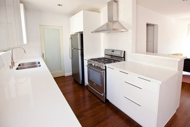 Example of a mid-sized trendy galley dark wood floor open concept kitchen design in Los Angeles with a double-bowl sink, flat-panel cabinets, white cabinets, solid surface countertops, white backsplash, subway tile backsplash, stainless steel appliances and no island