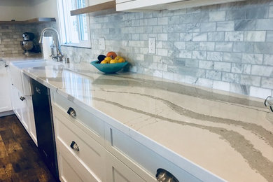 Example of a mid-sized trendy l-shaped vinyl floor and brown floor open concept kitchen design in Other with a farmhouse sink, shaker cabinets, white cabinets, quartz countertops, gray backsplash, marble backsplash, stainless steel appliances and an island