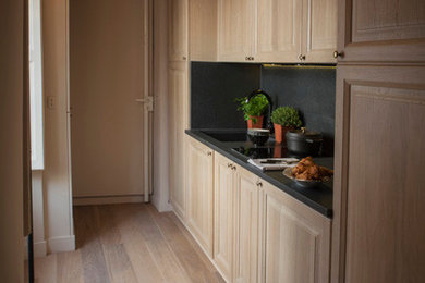 This is an example of a small transitional single-wall open plan kitchen in Paris with an integrated sink, beaded inset cabinets, light wood cabinets, granite benchtops, black splashback, limestone splashback, panelled appliances, light hardwood floors, no island, brown floor and black benchtop.