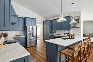 Modern inredning av ett stort vit vitt kök, med en undermonterad diskho, skåp i shakerstil, blå skåp, bänkskiva i kvarts, vitt stänkskydd, rostfria vitvaror, vinylgolv, en halv köksö och brunt golv