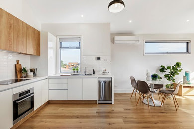 Design ideas for a contemporary l-shaped eat-in kitchen in Melbourne with a drop-in sink, flat-panel cabinets, white cabinets, white splashback, stainless steel appliances, medium hardwood floors, brown floor and white benchtop.