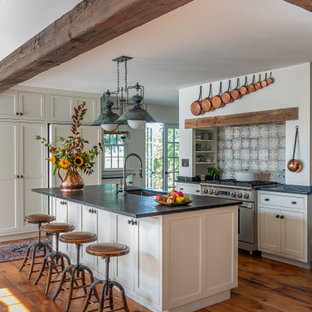 Farmhouse kitchen designs - Example of a farmhouse light wood floor and exposed beam kitchen design in Boston with white cabinets, stainless steel appliances, an island and black countertops
