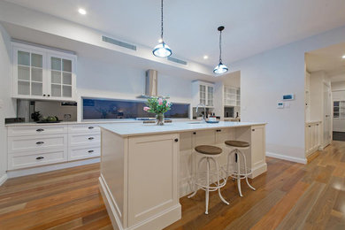 Photo of a large traditional galley open plan kitchen in Brisbane with an undermount sink, shaker cabinets, white cabinets, limestone benchtops, glass sheet splashback, stainless steel appliances, medium hardwood floors, with island, brown floor and white benchtop.