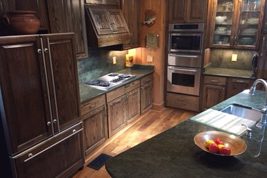 Large mountain style l-shaped medium tone wood floor and brown floor kitchen photo in Other with an undermount sink, raised-panel cabinets, dark wood cabinets, granite countertops, stainless steel appliances and an island