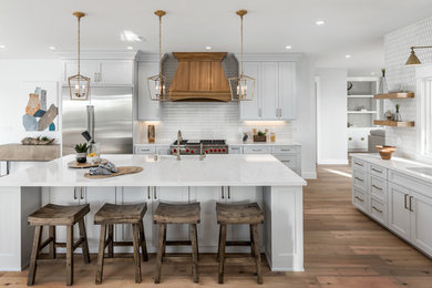 This is an example of a large country l-shaped eat-in kitchen in San Diego with white cabinets, white splashback, stainless steel appliances, medium hardwood floors, with island, brown floor, white benchtop, a farmhouse sink, shaker cabinets and quartz benchtops.