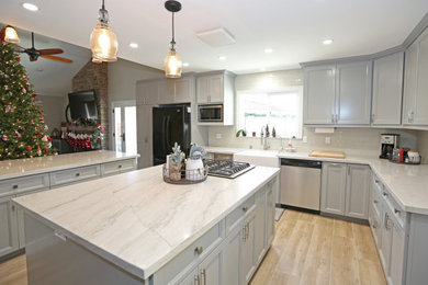 Example of a large l-shaped laminate floor and brown floor eat-in kitchen design in Orange County with a farmhouse sink, recessed-panel cabinets, gray cabinets, marble countertops, white backsplash, glass tile backsplash, black appliances, two islands and white countertops