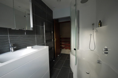 This is an example of a mid-sized modern master bathroom in Oxfordshire with an integrated sink, open cabinets, white cabinets, solid surface benchtops, a drop-in tub, a shower/bathtub combo, a wall-mount toilet, white tile, grey walls and slate floors.
