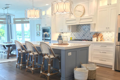 Mid-sized beach style medium tone wood floor and brown floor kitchen photo with a farmhouse sink, shaker cabinets, blue cabinets, quartz countertops, blue backsplash, glass tile backsplash, stainless steel appliances, an island and white countertops