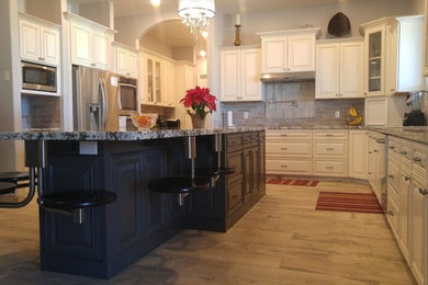 Example of a large classic l-shaped porcelain tile and brown floor eat-in kitchen design in Phoenix with an undermount sink, raised-panel cabinets, white cabinets, granite countertops, beige backsplash, stone tile backsplash, stainless steel appliances and an island