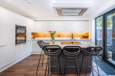 Photo of a medium sized contemporary l-shaped open plan kitchen in London with glass-front cabinets, white cabinets, quartz worktops, integrated appliances, an island, brown floors, white worktops, a submerged sink, orange splashback, glass tiled splashback and laminate floors.