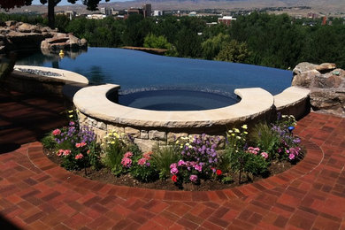 This is an example of a mid-sized modern backyard custom-shaped infinity pool in Boise with a hot tub and brick pavers.