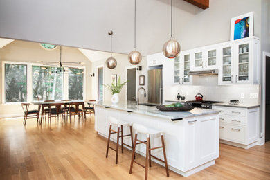 Example of a mid-sized transitional galley medium tone wood floor open concept kitchen design in New York with an undermount sink, shaker cabinets, white cabinets, granite countertops, white backsplash, ceramic backsplash, stainless steel appliances, an island and multicolored countertops