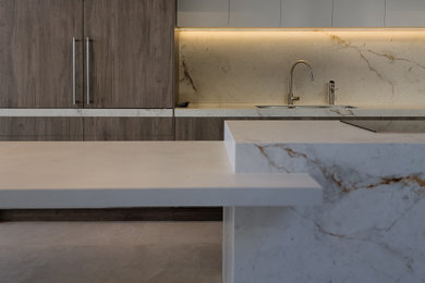 This is an example of a modern galley open plan kitchen in Melbourne with an undermount sink, flat-panel cabinets, medium wood cabinets, tile benchtops, white splashback, ceramic splashback, black appliances, ceramic floors, with island, beige floor and white benchtop.