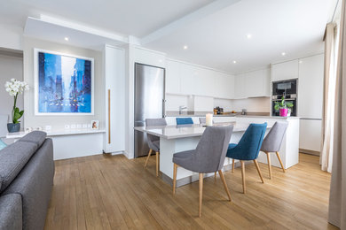Inspiration pour une cuisine américaine design en L de taille moyenne avec un évier encastré, un placard à porte affleurante, des portes de placard blanches, un plan de travail en quartz, une crédence blanche, une crédence en pierre calcaire, parquet clair, îlot et un plan de travail blanc.