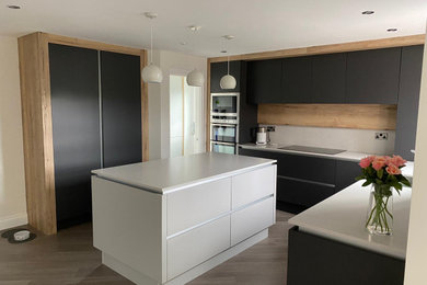This is an example of a medium sized contemporary u-shaped open plan kitchen in Devon with flat-panel cabinets, black cabinets, composite countertops, white splashback, an island, brown floors and white worktops.