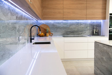 Photo of an expansive modern l-shaped eat-in kitchen in Newcastle - Maitland with an undermount sink, flat-panel cabinets, white cabinets, quartz benchtops, grey splashback, marble splashback, black appliances, porcelain floors, with island, grey floor and white benchtop.
