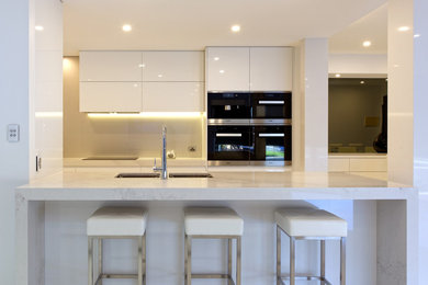 Photo of a mid-sized modern galley kitchen pantry in Melbourne with a double-bowl sink, white cabinets, quartz benchtops, white splashback, glass sheet splashback, black appliances and with island.